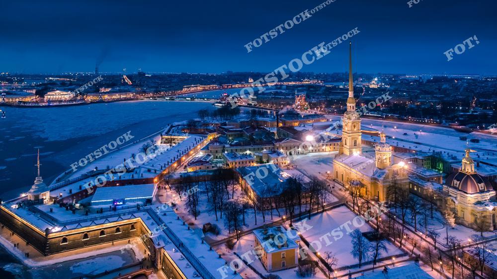 Панорамная фотография Петропавловской крепости и окружающего исторического центра Санкт-Петербурга, снятая с высоты птичьего полета в зимний вечер. На снимке видны крепостные стены, собор Петра и Павла, река Нева и освещенные улицы города. Съемка с дрона позволяет оценить масштаб архитектурных ансамблей, панораму Невы и величие Северной столицы России. Фотография передает атмосферу зимнего Петербурга, историческое наследие и уникальные панорамные виды города. Снимок идеально подходит для туристических публикаций, фотостоков, рекламы, оформления интерьеров и материалов о достопримечательностях Петербурга.